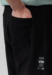 Schwarze Baumwollhosen mit einer seitlichen Tasche, sichtbaren Nähten und einem rechteckigen Markenetikett, auf dem weiße Schrift und Grafiken zu sehen sind.