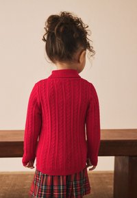 Piros kötött pulóver bordázott mintával és kis fonott mintákkal. Galléros nyakkivágással és hosszú ujjakkal, kockás szoknya fölött viselve.