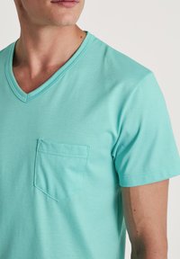 Una T-shirt corta a maniche con scollo a V di colore verde acqua, realizzata in tessuto morbido, con una tasca sul petto dai bordi cuciti e una texture liscia.