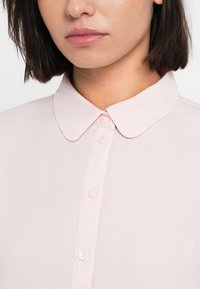 Ανοιχτό ροζ πουκάμισο με γιακά, κατασκευασμένο από υφάσμα με υφή, με σειρά από διακριτικά κουμπιά και κλασικό σχέδιο. Κοντινή λήψη της περιοχής του λαιμού.