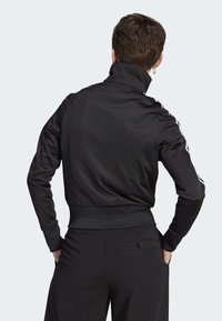 Chaqueta negra con cremallera, cuello alto, mangas largas y dobladillo acanalado. Presenta tres rayas blancas a lo largo de cada manga. Tejido suave y liso.