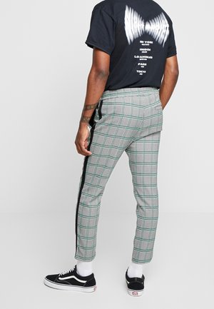 Mand iført sort t-shirt med grafik, ternede bukser med sorte sidelommer, hvide sokker og sorte sneakers, der står med ryggen mod kameraet.