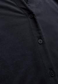 Camicia di tessuto nero con scollatura a V, caratterizzata da una superficie testurizzata e due bottoni neri sul davanti per la chiusura.