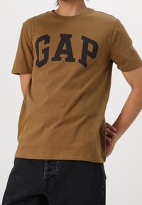 Camiseta de algodón marrón con el logotipo negro "GAP" impreso en la parte delantera. Cuello redondo clásico y mangas cortas. Textura suave y corte holgado.