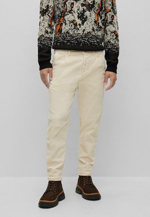 Man draagt beige corduroy broek, gebreide trui met zwart-oranje patroon en donkerbruine veterschoenen, staand tegen een effen achtergrond.