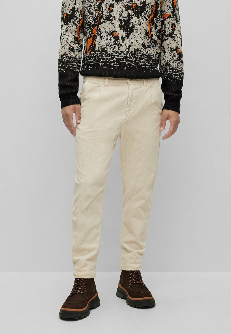 Homme portant un pantalon en velours côtelé beige, un pull à motifs noir et orange, et des bottines lacées marron foncé, debout devant un fond uni.