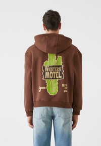 Ruda kapuca su dideliu žaliu kaktuso piešiniu ir tekstu ant nugaros. Pagaminta iš medvilnės, laisvas siluetas, ribotos krašteliai.