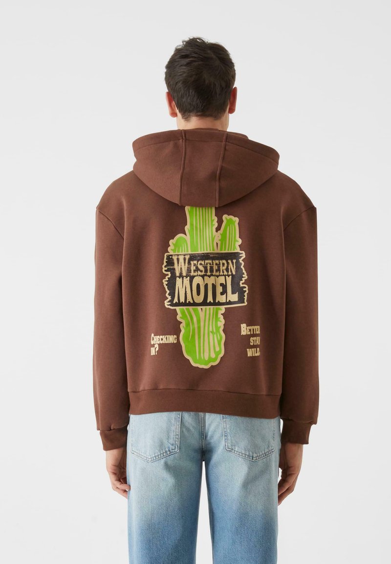 Ruda kapuca su dideliu žaliu kaktuso piešiniu ir tekstu ant nugaros. Pagaminta iš medvilnės, laisvas siluetas, ribotos krašteliai.