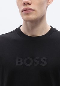 Fekete pamut pulóver, amelynek elején kiemelkedő fekete "BOSS" logó található, bordázott nyakkivágással és sima textúrával. Egyszerű és modern dizájn.