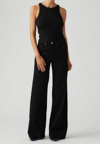 Débardeur noir côtelé associé à un jean noir taille haute à jambes larges. Le jean est doté de poches avant et d'une fermeture par bouton. Texture lisse sur toute la surface.