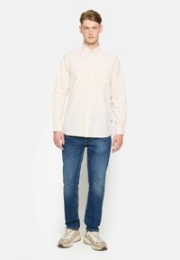 Helle beige gestreifte Knopfhemd, lange Ärmel, mit einer Brusttasche, kombiniert mit blauen Jeans und beigen Sneakers. Einheitlicher Hintergrund.