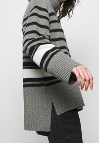 Szary sweter dzianinowy w pasy z czarno-białymi wzorami, z wysokim kołnierzem, prążkowanymi mankietami i bocznymi szczelinami, zestawiony z fakturowanymi czarnymi legginsami.