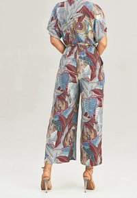 Mujer con un mono multicolor de estampado de hojas, con pantalones anchos y cintura ceñida con cinturón, combinado con sandalias de tacón alto transparentes, de espaldas.