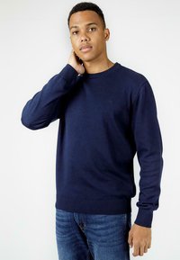 Granatowy, lekki sweatshirt z prążkowanymi mankietami i dołem, z okrągłym dekoltem i subtelnym logo na piersi. Noszony z niebieskimi dżinsami.