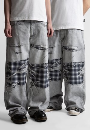 Dvaja ľudia oblečení v svetlosivých, výrazne opotrebovaných širokých džínsoch s čierno-bielymi károvanými záplatami a bielych košeliach.