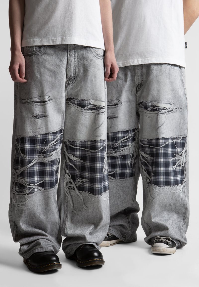 Δύο άτομα φορούν ανοιχτό γκρι, έντονα φθαρμένα παντελόνια με φαρδιά πόδια, διακοσμημένα με μαύρα και λευκά καρό μπαλώματα, και λευκά πουκάμισα.