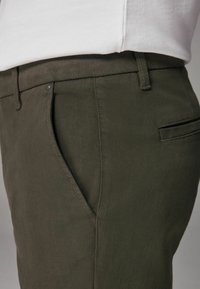 Pantalones verde oscuro con una tela texturizada, que presentan un bolsillo lateral, costuras sutiles y un bolsillo trasero con un diseño elegante.