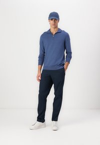 Men's blue zip-neck sweater in soft material, paired with dark navy pants and white sneakers. The cap is also blue, completing the look.