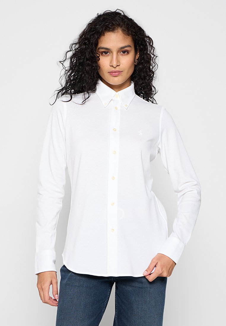 Camicia bianca a maniche lunghe con colletto classico e bottoni gialli. Presenta un logo discreto sul petto sinistro. Texture liscia.