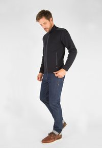 Veste en tricot noir avec fermeture éclair, col montant et deux poches zippées, associée à un jean en denim foncé et des chaussures à lacets marron.