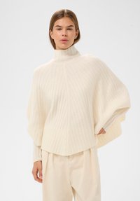 Kobieta ubrana w kremowy sweter ponczo z prążkowanym golfem i szerokimi rękawami, w połączeniu z kremowymi spodniami z wysokim stanem, stojąca na jednolitym tle.