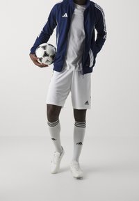 Marineblå Adidas träningsjacka, tillsammans med en vit t-shirt, vita shorts och strumpor med svarta ränder, som håller en svart och vit fotboll.