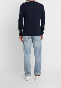 Marineblaues Langarmshirt mit weißen und grünen Seitenstreifen, kombiniert mit hellblauen Jeans und weißen Sneakers. Sichtbare Nähte und lockere Passform.