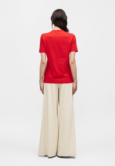 Άτομο με μακριά σκούρα μαλλιά που φορά κόκκινο κοντομάνικο πουκάμισο και μπεζ παντελόνια με φαρδιά μπατζάκια, στέκεται με την πλάτη προς ένα απλό φόντο.