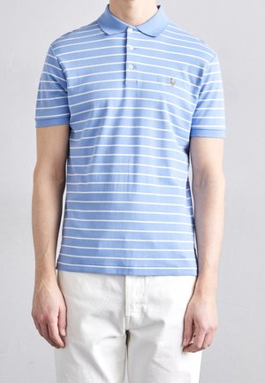 Man draagt een lichtblauw-wit horizontaal gestreept poloshirt met een witte broek, staand tegen een effen lichtgrijze achtergrond.