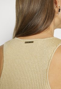 Vaaleankultainen ribattu toppi pyöreällä kaula-aukoalla, jossa on pieni musta ja kultainen brändilappu takana. Pehmeä pinta ja napakka malli.