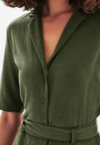 Combinaison culotte vert olive, à manches courtes, avec col, fermeture à boutons sur le devant et une ceinture en tissu à la taille. Texture douce avec une finition unie.