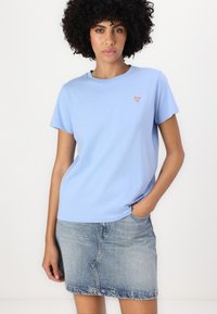 T-șirt de bumbac de culoare albastru deschis, cu mâneci scurte și guler rotund, având un mic logo în formă de triunghi roșu pe pieptul stâng. Purtați cu o fustă din denim.