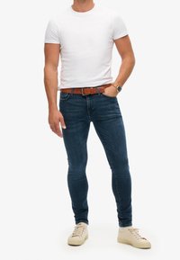 Blå slim-fit jeans med blekta detaljer, bärs med en vit t-shirt med kort ärm och ett brunt läderbälte. Beiga sneakers kompletterar outfiten.
