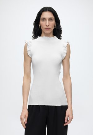 Femme aux cheveux bruns ondulés, vêtue d'un haut blanc sans manches avec des bords volantés et d’un pantalon noir, face à un fond uni.
