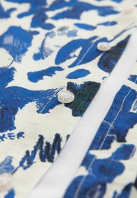 Tela de camisa de patrón azul y blanco con diseños florales abstractos. Presenta un botón blanco con el texto "Ted Baker" y detalles de costura visibles.