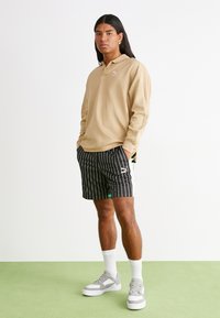 Beżowa koszulka polo z długim rękawem i kołnierzykiem, zestawiona z czarno-białymi spodenki w paski. Szare sneakersy z grubą białą podeszwą.