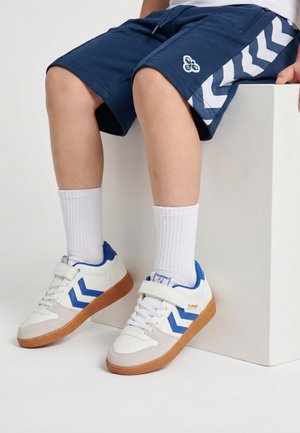 Dziecko siedzi na białym bloku, nosi granatowe spodenki z białymi pasami w jodełkę oraz białe trampki z niebieskimi akcentami i gumowymi podeszwami.