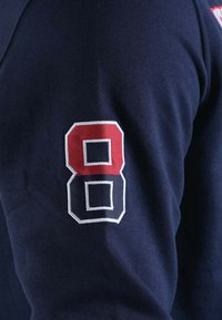 Chemise à manches longues bleu marine avec un grand numéro 8 brodé en rouge et blanc sur la manche droite, fabriquée en tissu doux avec une texture lisse.