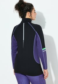 Žena s kudrnatými vlasy staženými do vysokého culíku, oblečená v černém a fialovém atletickém topu s dlouhým rukávem a fialových legínách s vzorem, čelící bílé zdi.