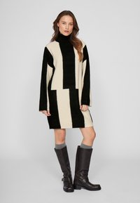 Adīta kleita ar augsto apkakli melnā un krēmkrāsā, ar vertikālām un horizontālām svītrām. Valkāta ar ceļa garuma brūnām zābakām.