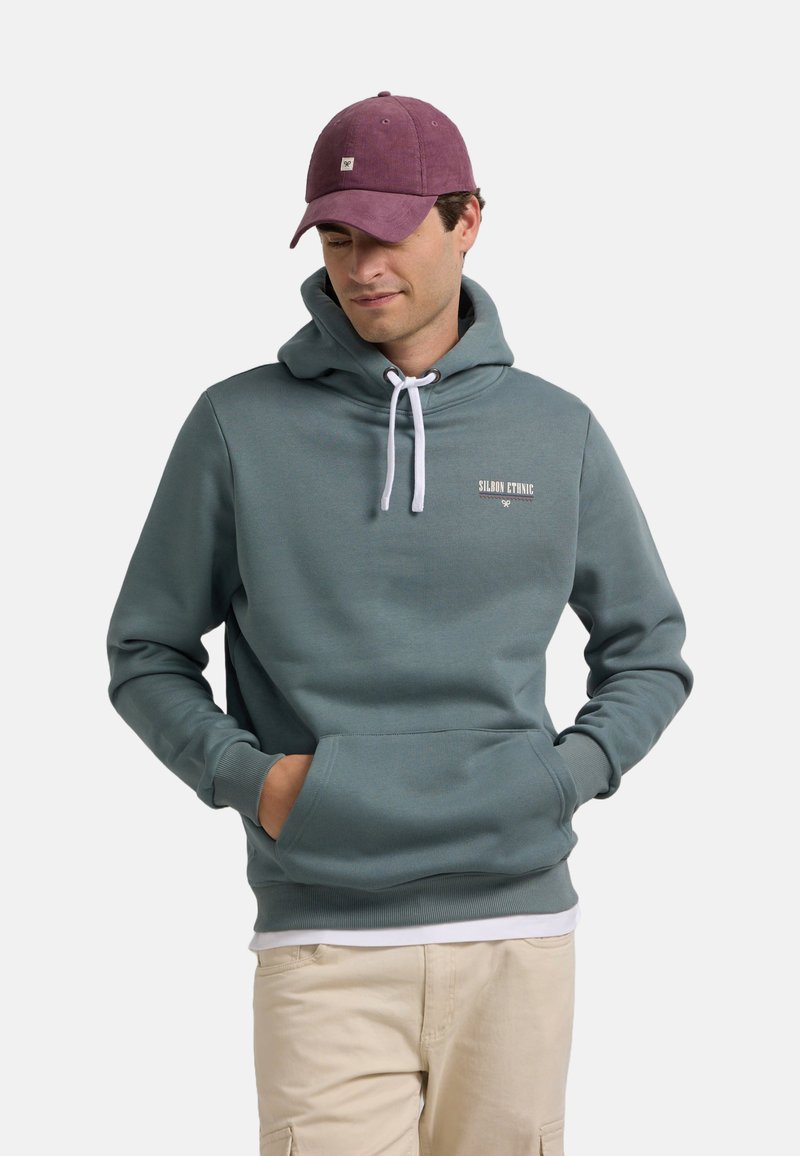 Sudadera con capucha en azul verdoso apagado, con un bolsillo canguro, cordones blancos y un sutil detalle de logo. Combinada con una gorra burdeos.