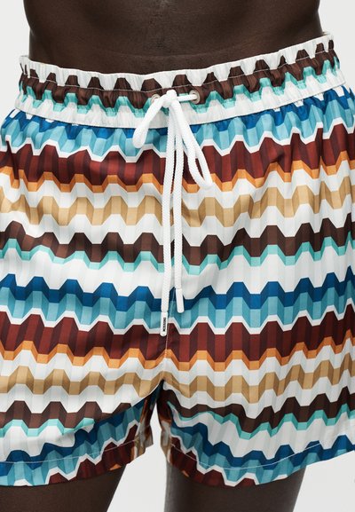 Short de bain multicolore à motif zigzag avec cordon blanc porté par une personne à la peau foncée, visible de la taille jusqu'à mi-cuisse.