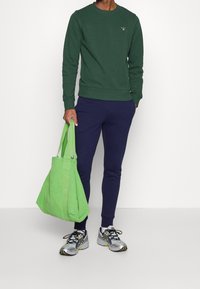 Person som bär en mörkgrön sweatshirt och marinblå joggers, håller en stor ljusgrön tote-väska, med vita och silverfärgade löparskor.