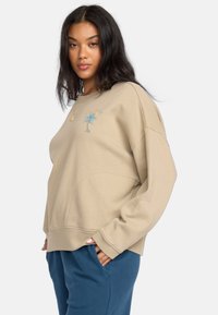 Beżowy sweter z luźnym krojem, z niebieską i zieloną haftowaną palmą na piersi. Miękka tkanina i ściągacze.