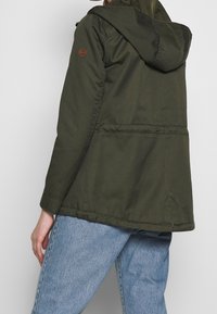 Kurtka w kolorze oliwkowej zieleni z kapturem, wykonana z gładkiego materiału. Posiada naszywkę z logo na rękawie oraz średniej długości fason.