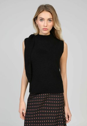 Femme blonde portant un pull sans manches noir avec un détail écharpe et une jupe noire à pois marron, debout devant un fond gris.