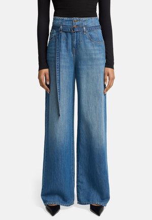 Vrouw draagt hooggetailleerde wijde blauwe jeans met aangehechte denimriem en zwarte schoenen met puntige neus, gecombineerd met een zwart top met lange mouwen.