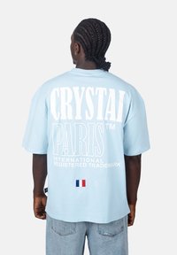 Světle modré oversized tričko vyrobené z bavlny. Na zadní straně se nachází velký bílý nápis "CRYSTAL PARIS" a malý detail francouzské vlajky.