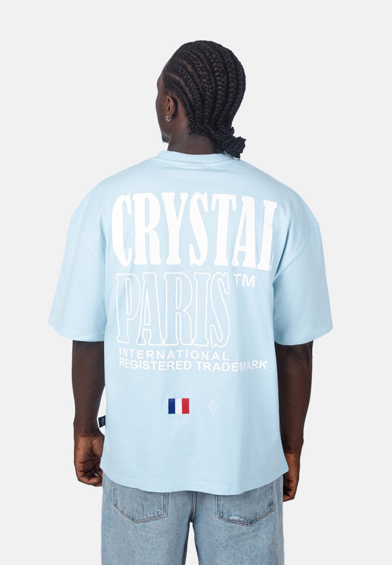 Světle modré oversized tričko vyrobené z bavlny. Na zadní straně se nachází velký bílý nápis "CRYSTAL PARIS" a malý detail francouzské vlajky.