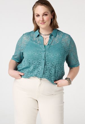 Vrouw met kort haar, draagt een blauwgroen kanten kort mouwen bloesje met knopen over een tanktop en crème broek, handen in de zakken, licht glimlachend.